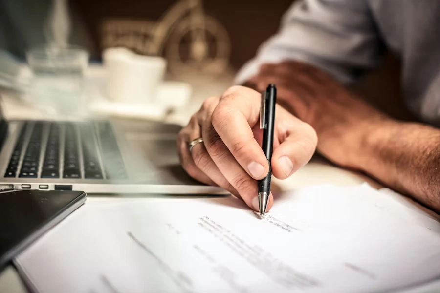 Person signing a document at a desk with a laptop and smartphone nearby