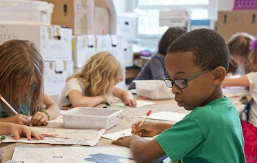 A young boy with glasses drawing at a desk in a classroom sitting with other children